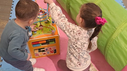 Two young children playing with a wooden toy on pink and blue mats with a colourful play tunnel next to them.